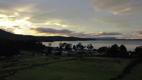 Vista-Aérea-Lenta-De-Un-Dron-Sobre-Llanuras-De-árboles-Y-Pasto,-Puesta-De-Sol-Lenta-En-La-Playa-De-La-Isla-Al-Anochecer,-Cielo-épico-Y-Cordillera-Con-Pequeñas-Chozas-Y-Bosque-Con-Nubes