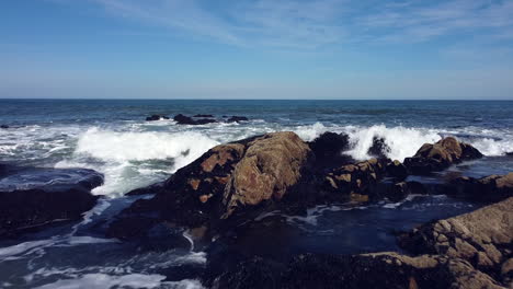 Drone-flying-from-beach-to-ocean-over-waves-breaking-on-rocks