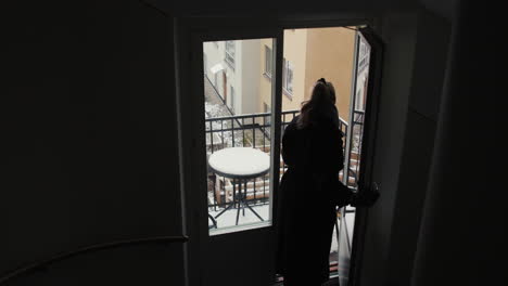 Blonde-young-woman-in-black-coat-going-out-on-snowy-apartment-balcony