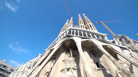 Vista-Amplia-Y-Suave-Hacia-Arriba-De-La-Fachada-De-Cristo-Con-Cielo-Azul-En-La-Sagrada-Familia-Con-Grúas-De-Construcción,-Barcelona