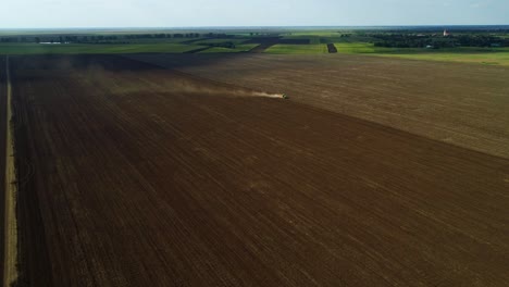 Aerial-push-in-shot-of-a-green-tractor-seeding,-sowing-agricultural-crops-at-the-field-on-a-sunny-day-with-blue-sky-and-white-clouds