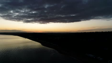 Drone-Con-Vista-Al-Agua-Tranquila-Y-Brillante-Al-Amanecer-Sobre-Un-Paisaje-De-Silueta-Oscura-Y-Nubes