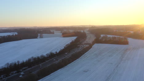 Drone-footage-of-motorway-surrounded-by-snow-in-fields-with-cars-driving-along-the-M25-winter-morning-for-sunrise