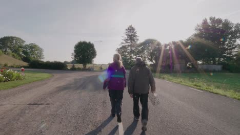 Una-Pareja-De-Ancianos-Camina-Por-Un-Camino-Rural-Con-Una-Bolsa-Blanca-En-Un-Pequeño-Pueblo,-Plano-General-Que-Sigue-Hacia-Adelante