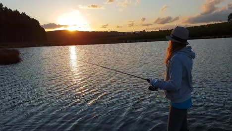 Girl-fishing-from-a-pier-in-a-calm-lake-with-the-sun-setting-in-the-distance