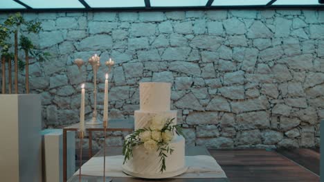 three-tiered-wedding-cake-decorated-with-white-flowers,-placed-on-a-table-against-a-rustic-stone-wall,-accompanied-by-tall-candles