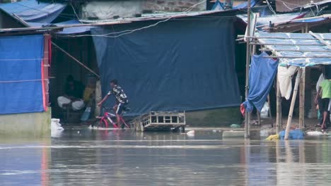 Young-Asian-boy-rides-a-bicycle-in-flood-water-of-a-slum-as-muddy-water-flows-in-the-street