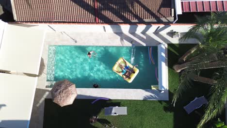 Friends-at-swimming-pool-aerial-drone-view-of-people-that-swim-at-pool-floating-over-inflatable-bed-in-chalet-at-residential-area