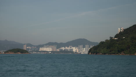 Vista-Del-Puerto-De-Hong-Kong-Desde-Un-Ferry-En-Movimiento-Con-Laderas-Verdes-Y-Edificios-Densos-Durante-El-Día