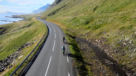 Un-Dron-Sigue-A-Personas-En-Bicicleta-En-Una-Curva-De-Una-Carretera-En-Islandia-Sobre-El-Océano-En-Dirección-A-Las-Montañas