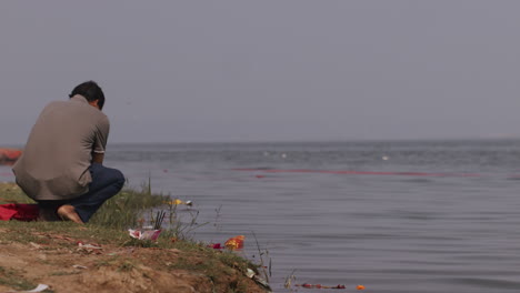 A-hand-held-long-shot-of-an-Indian-man-sitting-with-his-back-towards-the-camera-on-the-banks-of-a-river-and-doing-the-last-rites,-as-per-Hindu-religion,-of-a-dead-person-by-throwing-ashes-in-water