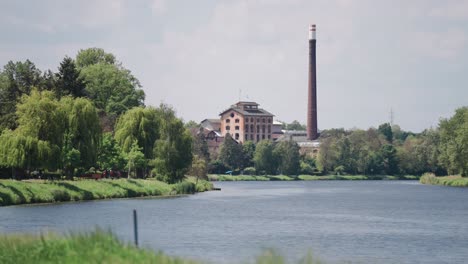 Industrial-building-with-a-tall-chimney-next-to-a-river,-surrounded-by-lush-green-trees