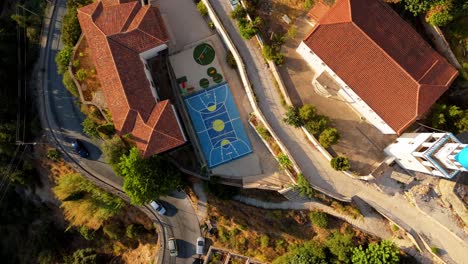 A-hillside-village-with-a-vibrant-blue-basketball-court-in-the-center,-aerial-view