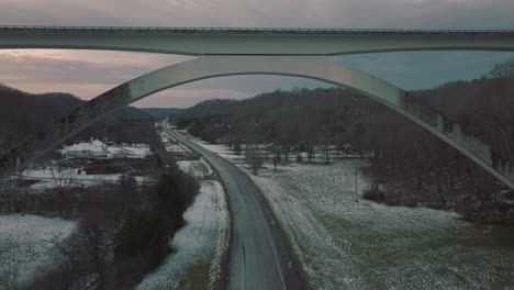 Flying-along-country-road-running-under-Natchez-Trace-Bridge-at-dusk-in-winter,-landscape-covered-in-light-snow