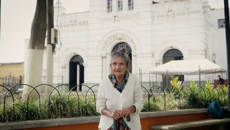 Elderly-woman-with-traditional-attire-stands-calmly-before-a-historic-church-in-Santa-Barbara,-Antioquia,-showcasing-cultural-heritage-and-peaceful-surroundings