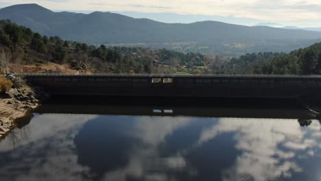 Drone-flight-over-a-dam-in-the-Tiétar-valley-where-we-see-the-reflection-of-the-sky-in-the-water-and-passing-the-retaining-wall-we-see-the-work-of-a-road,-the-valley-appears-in-the-background