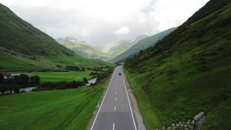 Road-in-Swiss-Alps-with-small-traffic-car-and-rainbow-at-the-sky-shot-from-a-drone