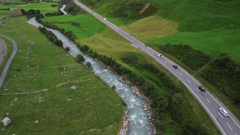 Traffic-on-a-road-in-Swiss-Alps-with-cars,-cyclists-andsmall-stream-river-shot-from-a-drone