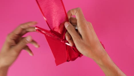 Atar-Un-Lazo-De-Cinta-Roja-Alrededor-De-Una-Caja-De-Regalo-Para-San-Valentín,-Fondo-Rosa