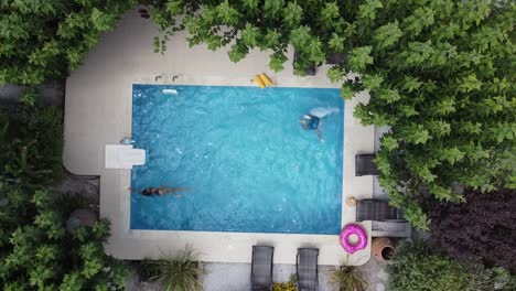 Young-couple-enjoying-summer-by-pool---both-swimming-in-pool---drone-hovering-over-pool-in-bird's-eye-view