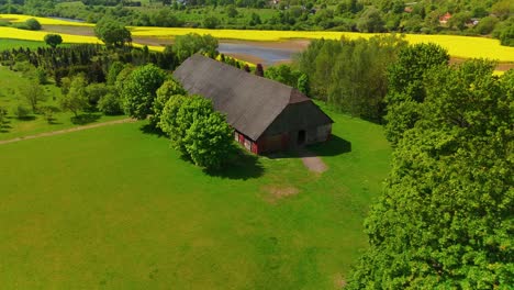 Aerial-view-of-a-rustic-barn-surrounded-by-lush-green-fields,-trees,-and-distant-yellow-crop-fields