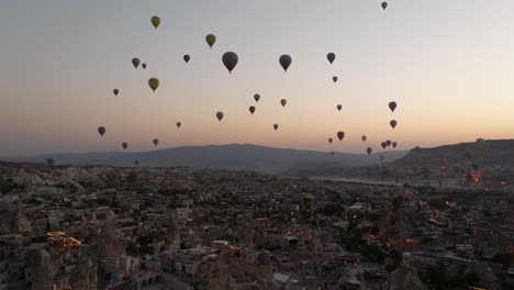 Drone-ascends-gradually-past-stone-spire-towers-with-green-glow-on-horizon-against-silhouette-of-hot-air-balloons,-Cappadocia-Turkey