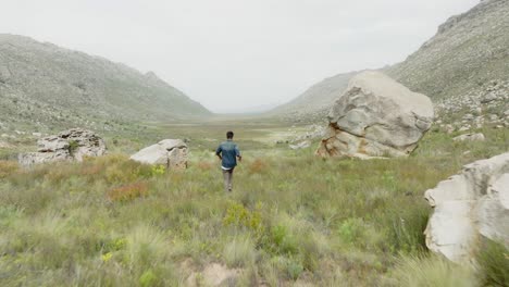 Un-Joven-Camina-En-Un-Paisaje-Alto-Junto-A-Una-Gran-Roca-En-El-área-Silvestre-De-Cederberg-En-Sudáfrica---Un-Dron-Volando-Detrás-De-él