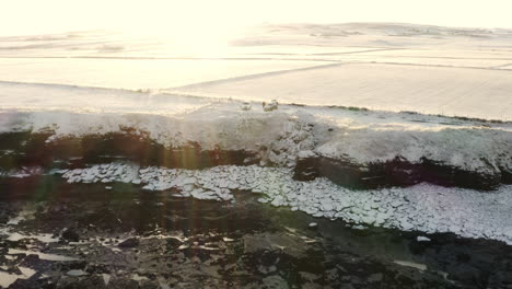 Drone-descends-as-sun-flare-spreads-light-across-surfers-hanging-out-at-top-of-sea-cliff-by-cars-on-cold-snowy-winter-day