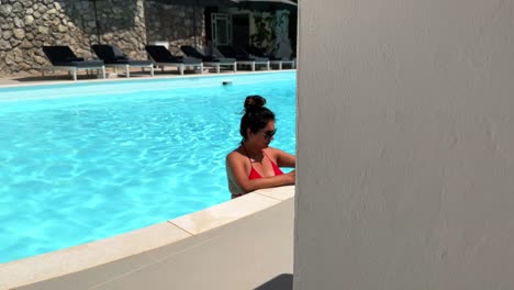 Camera-peeks-around-wall-to-reveal-brunette-woman-relaxing-in-pool-reading-book-enjoying-vacation