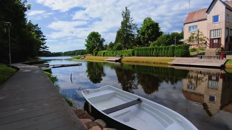 A-tranquil-lakeside-scene-featuring-a-white-rowboat-tied-to-a-dock,-a-wooden-house,-and-calm-water-reflecting-the-surrounding-greenery