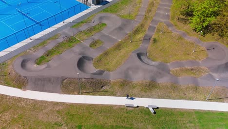 Aerial-view-of-a-curvy,-paved-bike-and-skate-track-in-a-park,-surrounded-by-greenery-and-adjacent-to-a-tennis-court