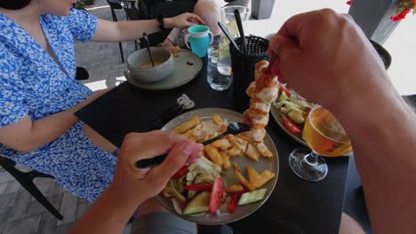 Una-Persona-Disfruta-De-Una-Comida-De-Brochetas-De-Pollo-A-La-Parrilla-Con-Papas-Fritas-Y-Verduras-Frescas-En-Un-Café-Al-Aire-Libre.