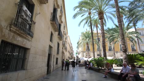 A-bustling-city-street-in-Alicante-shows-people-resting-along-the-side-of-a-large-building,-enjoying-the-relaxed-atmosphere-and-vibrant-urban-surroundings