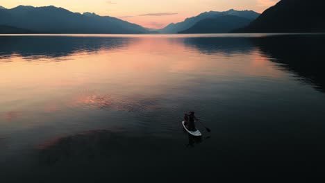 Couple-sit-on-an-inflatable-paddle-board-as-they-float-and-paddle-around-the-huge-Harrison-Lake-in-Chilliwack-British-Columbia-at-a-golden-hour-sunset-with-orange-reflections-on-the-water