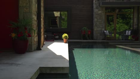 Side-to-side-movement-across-a-long-luxury-blue-infinity-swimming-pool-in-back-yard-on-sunny-summer-day-surrounding-a-property-detailed-with-stone,-wood-and-brick