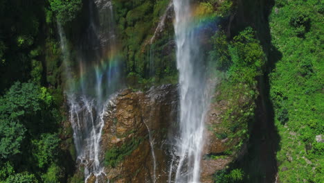 Drone-shot-of-huge-waterfall-and-rainbow-over-lush-greenery-in-Nepal-stunning-view-shows-a-massive-cliff,-offering-exploration-fun-and-adventurous-travel-to-Nepal’s-authentic-natural-areas