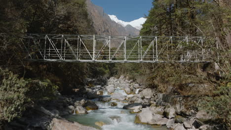 Drone-shot-captures-a-trekker-walking-on-bridge-in-Langtang-National-Park-Nepal-adventurer-admires-wilderness-and-mountainous-landscape-rivers,-trees,-and-the-bright-Langtang-Himalayas-in-backdrop