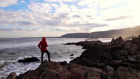 Man-stands-on-Hermanus-coastline-vantage-point-at-sunset,-drone-fly-by