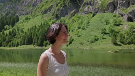 A-smiling-woman-walks-near-a-peaceful-lake-with-lush-mountains-and-trees-in-the-background