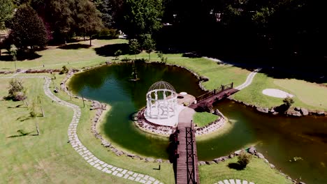 Aerial-view-of-a-picturesque-lake-with-a-white-gazebo-and-wooden-bridge-on-a-manicured-lawn