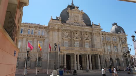 A-front-view-of-the-majestic-City-Hall-building,-or-Palacio-Consistorial,-with-people-walking-back-and-forth,-capturing-the-lively-atmosphere-of-the-area