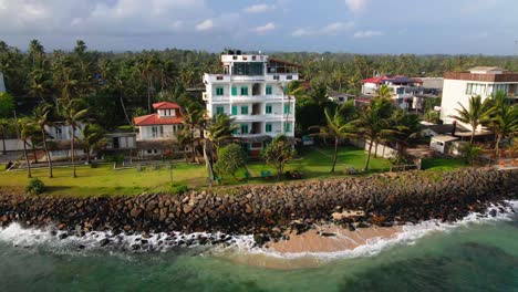 Aerial-view-of-a-coastal-hotel-by-the-ocean-in-Ahangama,-Sri-Lanka,-featuring-palm-trees,-a-rocky-shore,-and-surrounding-buildings