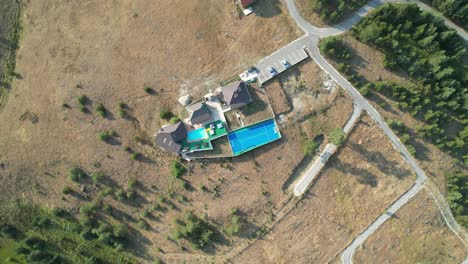 Aerial-perspective-showing-several-houses-with-a-pool-and-sports-court,-located-in-an-expansive,-dry-countryside-area,-surrounded-by-sparse-vegetation-and-winding-roads