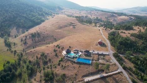 Aerial-view-of-a-picturesque-hillside-in-Zlatibor,-Sainovci,-Serbia,-featuring-a-poolside-resort-nestled-in-a-lush,-hilly-landscape,-surrounded-by-rolling-fields,-trees,-and-serene-rural-roads