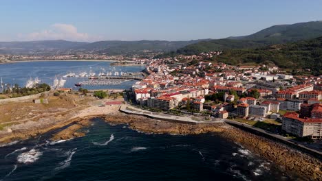 aerial---bayona-village-with-coastline,-marina,-red-roofed-buildings,-and-mountainous-backdrop