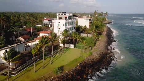 Aerial-footage-of-a-hotel-along-the-coast-of-Ahangama,-Sri-Lanka,-showing-a-lush-landscape-with-palm-trees-and-ocean-waves-hitting-the-rocky-shoreline