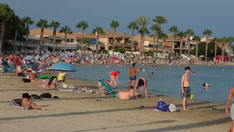 People-relax-in-the-sunshine-and-enjoy-the-cool-breeze-at-Los-Alcázares-beach,-soaking-up-the-summer-vibe,-swimming,-and-unwinding-by-the-sea