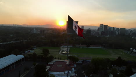 Drone-circling-a-mexican-flag-with-evening-sunburst-background,-in-Mexico-city