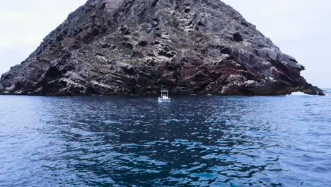 Aerial-pullback-from-boat-anchored-below-rocky-island-in-Coronado-Islands,-Mexico,-surrounded-by-the-vast-blue-ocean,-centered-fishing-trip