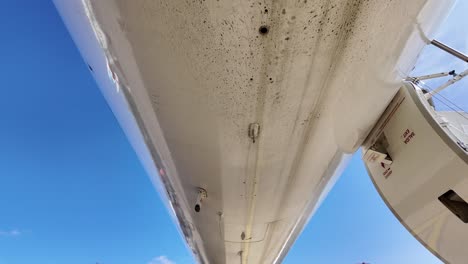 External-view-of-the-lower-part-of-the-fuselage-of-a-dirty-white-color-fuselage-jet-from-the-nose-to-thre-rear-part,-tilting-down,-with-some-buildings-at-the-back